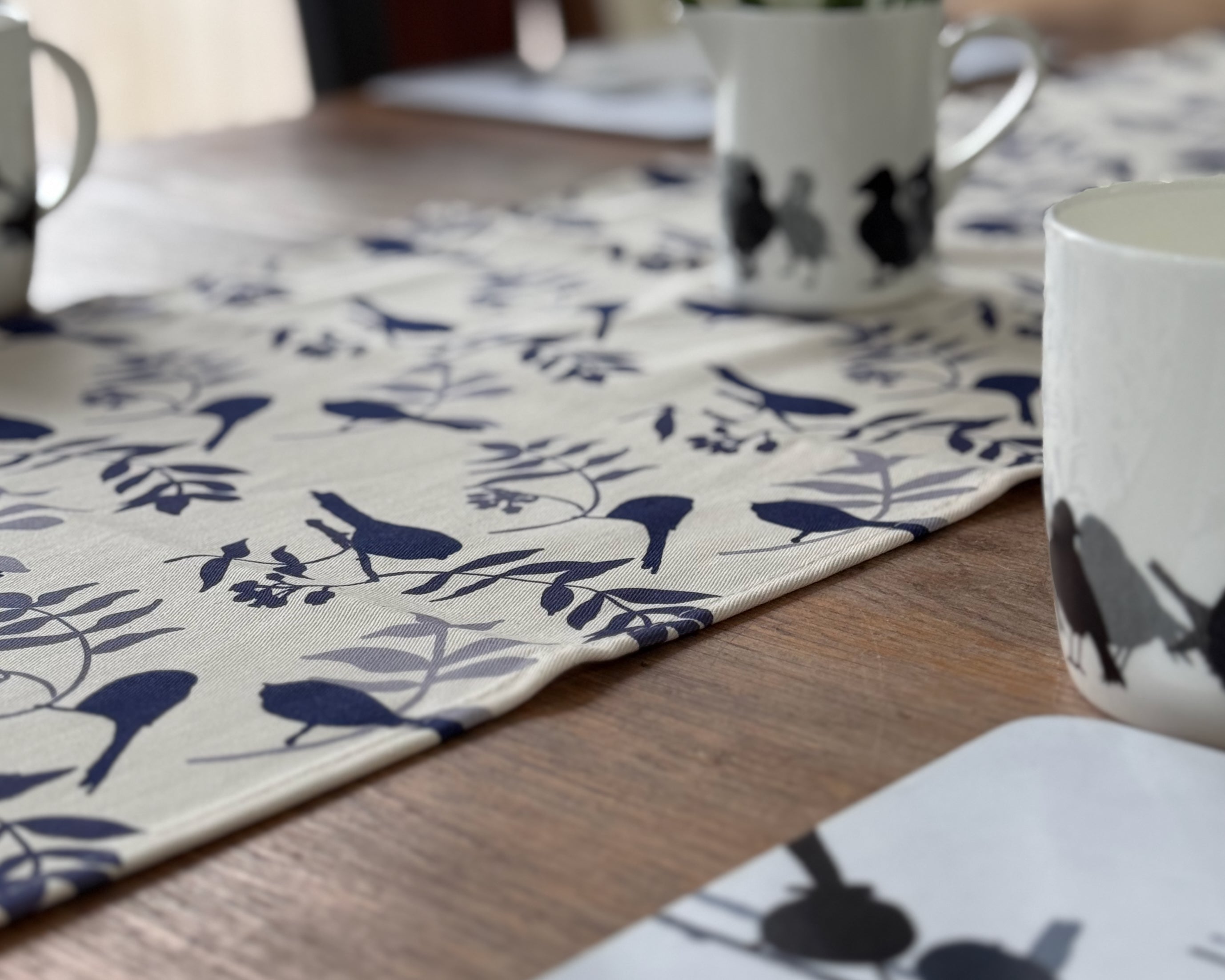 Table setting with white mugs and placemats featuring blue bird patterns on a wooden table.